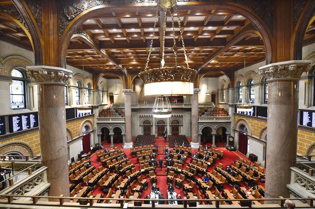 A view of the ornate NY State Assembly chamber, which has a soaring ceiling, a red carpet and pillars
